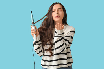 Beautiful young woman with curling iron blowing kiss on blue background