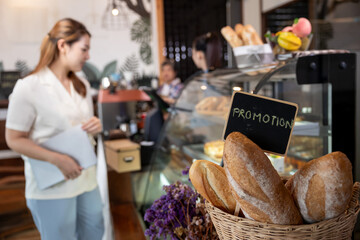 Freshly baked bread with promotion sign in basket placed near cafe counter. Asian young woman standing holding tablet while observing display shelf. Warm bakery ambiance indoor