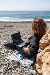 Beach Laptop Woman: Girl uses laptop on sunny pebble beach, next to the rock, for remote work.