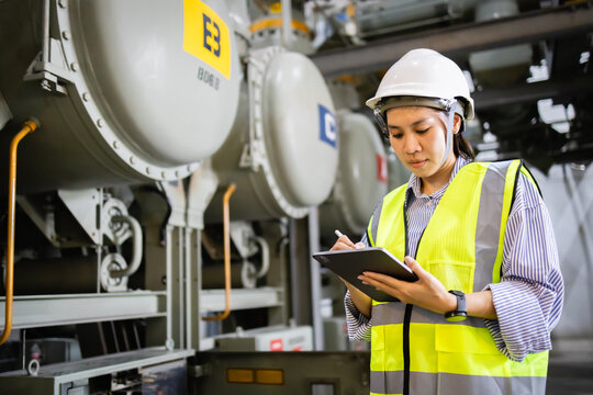 Female engineer wearing safety helmet and insulated vest inspects electric grid switchgear in field while taking notes on clipboard near gas insulated switchgear equipment