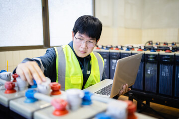 Technician wearing safety vest inspects backup power system battery while using laptop to ensure proper maintenance and reliable energy supply in industrial setting