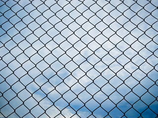A close-up view of a metal mesh fence with a backdrop of a blue sky and scattered clouds. 