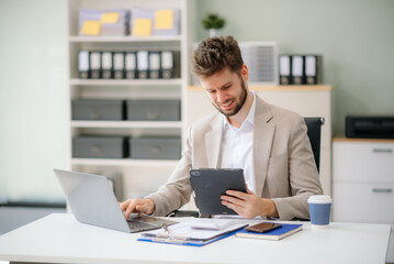 Confident businessman working on finance analysis with a tablet and laptop at office desk.