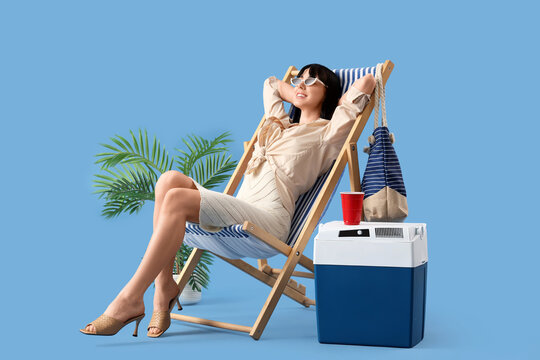 Relaxing woman with portable fridge on beach chair against blue background - Powered by Adobe