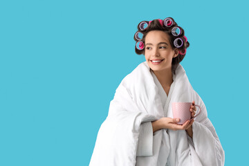 Young woman in blanket with hair curlers and coffee cup on blue background