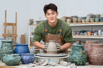 Young man is shaping clay pot on pottery wheel in bright studio filled with various ceramic pieces. His focused expression reflects his passion for pottery