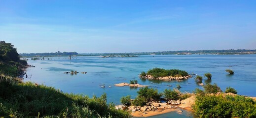 A wide panoramic view of the vast Mekong River under a clear blue sky. The river flows past lush green banks and small rocky islands, with distant trees outlining the horizon.
