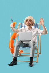 Young businessman with laptop and cocktail waving hand in deck chair on blue background. Summer...
