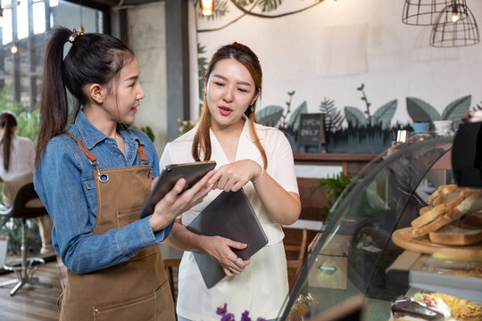 Asian young woman entrepreneur in white blouse holding tablet discussing plan with female staff in apron using digital tablet inside cozy bakery shop. Collaboration and small business teamwork