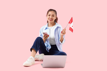 Young Asian woman with flag of Canada using mobile phone and laptop on pink background