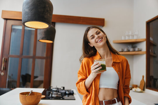 Young woman in an orange shirt holding a green smoothie in a modern kitchen, showcasing healthy living and happiness with a bright, inviting atmosphere