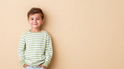 A cheerful young boy wearing a green and white striped shirt stands against a beige background, radiating happiness and confidence. The image includes ample copy space for versatile usage.