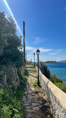 Stone Pathway With Vintage Street Lamps Along The Seaside On A Sunny Day, With Coastal Vegetation And Blue Water, Concept Of Tranquility, Travel, And Summer Exploration