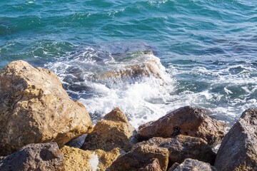 Rocky shore with turquoise sea water rolling in waves and creating splashes and thick white sea...