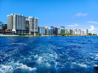 Naklejka premium Malé cityscape stretches along the horizon, showcasing numerous buildings along the coastline under a blue sky, taken from the water with a visible boat wake in the foreground. Embodies urban travel