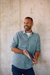Happy entrepreneur with laptop leaning on wall and looking away.