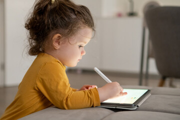 Small girl using a stylus to create artwork on a tablet. Living room setting with natural light and...