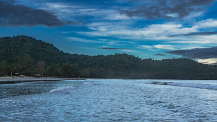 Morning seascape. The ocean waves are foaming and spreading. Sun loungers, umbrellas, and straw awnings are visible on the sandy beach. A green hill against a blue sky and clouds. Malaysia.Borneo.