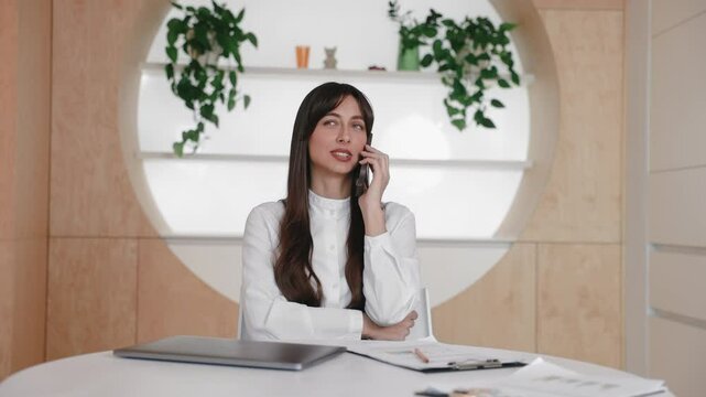 Serious stylish Caucasian female sitting at modern desk and talking on smartphone while crossing arms. Discussing project or handling business call. Solving issue or negotiating details remotely.