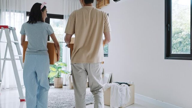 Young Asian couple working together, carrying cardboard boxes while moving into their new house. Woman walking upstairs and man entering empty room, starting a new chapter of life with teamwork.
