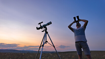 Amateur astronomer using telescope and binoculars for watching stars, Moon, planets and other celestial objects from a countryside field in nature.