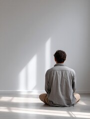 Young caucasian male sitting alone in minimalist room facing blank wall.