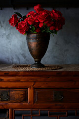 A rustic bronze vase filled with vibrant red roses is displayed on a wooden table againt a textured...