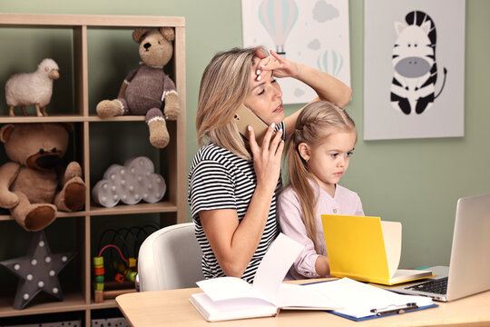 Work-family balance. Overwhelmed single mother talking by smartphone and her daughter at table in child's room
