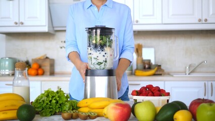 Woman using a blender, preparing a healthy smoothie with various fruits and vegetables like spinach, banana, kiwi, strawberry, apple, avocado in a bright kitchen