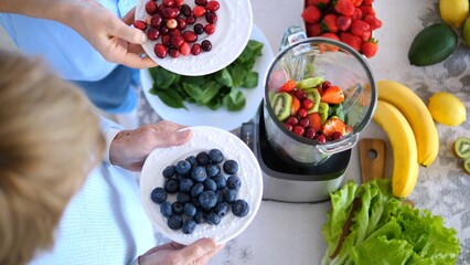 Two women holding plates with blueberries and cranberries, preparing ingredients for a healthy smoothie with blender, fruits and vegetables on kitchen table