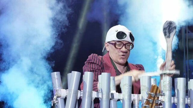 A clown in a funny costume entertains guests in the circus arena in the light of colorful spotlights and smoke. A clown plays a musical instrument in the light of colorful lights. Circus performance.