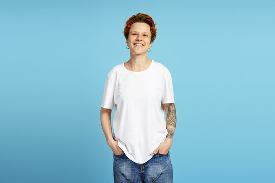 Young person with orange hair and tattoos smiling with hands in pockets - Powered by Adobe