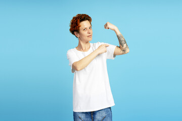 Young person showing strength and pointing at biceps with tattoo on blue background