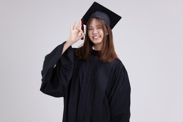 College degree graduations. Happy young Asian girl in gown with mortarboard shows OK hand sign...