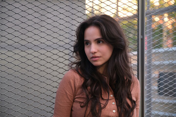Portrait of a young Latin woman with long brown hair, wearing a light brown shirt, standing near a metal mesh fence in an urban environment