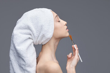 Young caucasian female applying makeup with brush in towel turban against gray background.