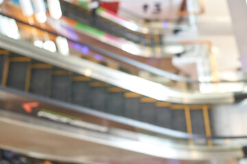 Blurry view of escalators in shopping mall with colorful lights and reflections, featuring busy modern architecture.
