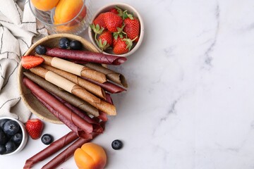 Tasty fruit leather rolls, apricots and berries on white marble table, flat lay. Space for text