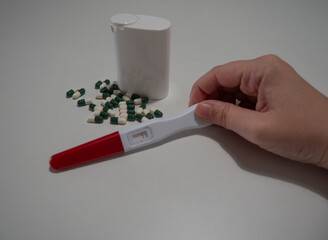 Close-up of a hand holding a pregnancy test with a red and white handle, next to a group of green and white capsules and a white pill container on a plain white background, representing health