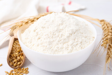Wheat flour in bowl, grains and spikes on white table, closeup