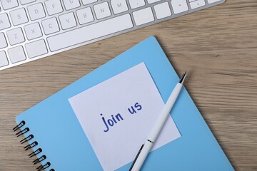 Sticky note with phrase Join Us, pen, notebook and computer keyboard on wooden table, flat lay