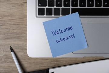 Sticky note with phrase Welcome Aboard, laptop and pen on wooden table, flat lay