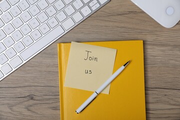Sticky note with phrase Join Us, pen, notebook, computer keyboard and mouse on wooden table, flat...