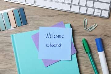 Paper note with phrase Welcome Aboard, keyboard and stationery on wooden table, flat lay