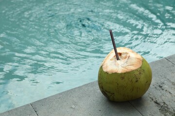 Green coconut with refreshing drink and straw near swimming pool outdoors, space for text