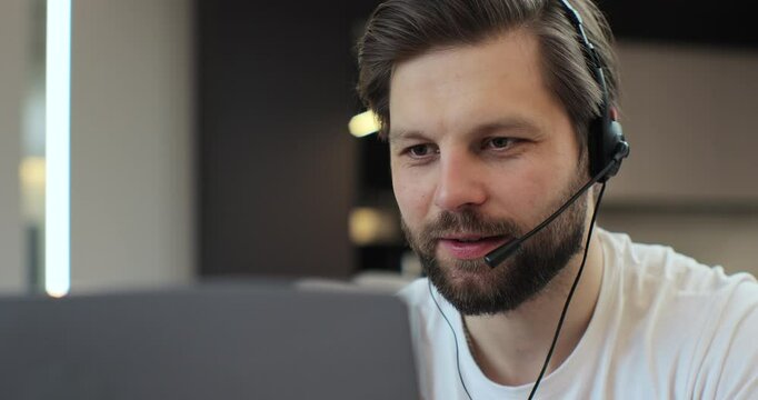 Closeup face man wearing headset and talking, call center worker. Positive young man wearing headset. Caucasian man holding meeting with colleagues in home interior.