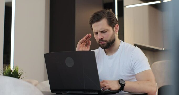 Tired man is working on laptop computer at the desk in kitchen at home office. Man upset stressed working at the laptop reading bad news. Concept of remote work or education.