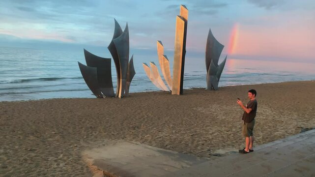 A person photographs the Les Braves monument by Anilore Banon on Omaha Beach, Normandy, at sunset, with rainbow over the sea, WWII memorial sculpture in stainless steel, soft light, camera static