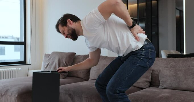 Young man suffering from back pain sitting on sofa in living room at home. Man holds lower back with hands, massages sore muscles. People health care problem.