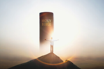 Christian standing with arms raised on top of a high mountain, praising and praying, looking at the holy bible and the cross of Jesus Christ with love in the background
